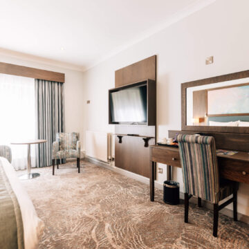 Spacious hotel room with cosy seating, large window, desk, and wall-mounted TV, offering a relaxing stay.