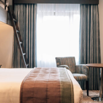 Cosy hotel room with a double bed, striped duvet, chairs, and natural light through large window with curtains.