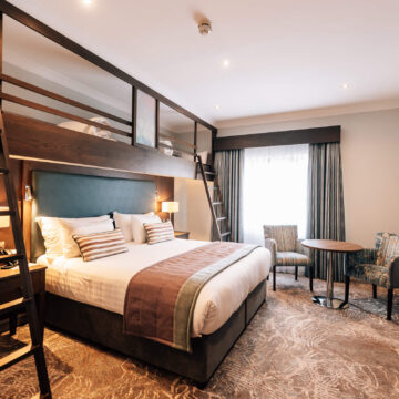 Stylish hotel room with plush double bed, loft bunk bed, cosy seating area, flat-screen TV, and modern decor.