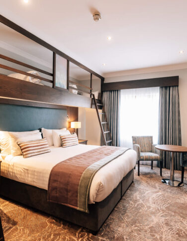 Stylish hotel room with plush double bed, loft bunk bed, cosy seating area, flat-screen TV, and modern decor.