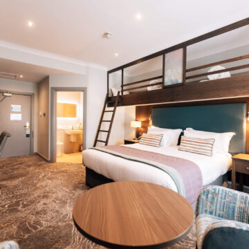 Spacious hotel room with cosy double bed, modern decor, loft-style bunk bed and ensuite bathroom, ideal for families.