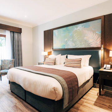 Elegant hotel room with king-size bed, modern decor, artwork, wooden flooring, and ample natural light.