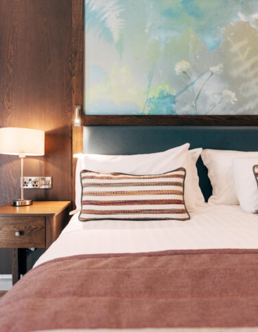 Cosy hotel room with plush bed, elegant wooden decor, bedside lamp, and tranquil nature artwork, promising a restful stay.