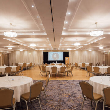 Elegant hotel conference room with round tables, plush chairs, crystal chandeliers, and a large projector screen.
